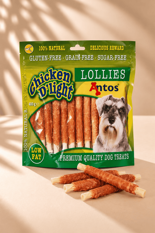Antos Chicken D'Light Lollies 400 g, groen-gele zak met kipkauwstaafjes, hondenafbeelding en losse snacks op beige achtergrond.