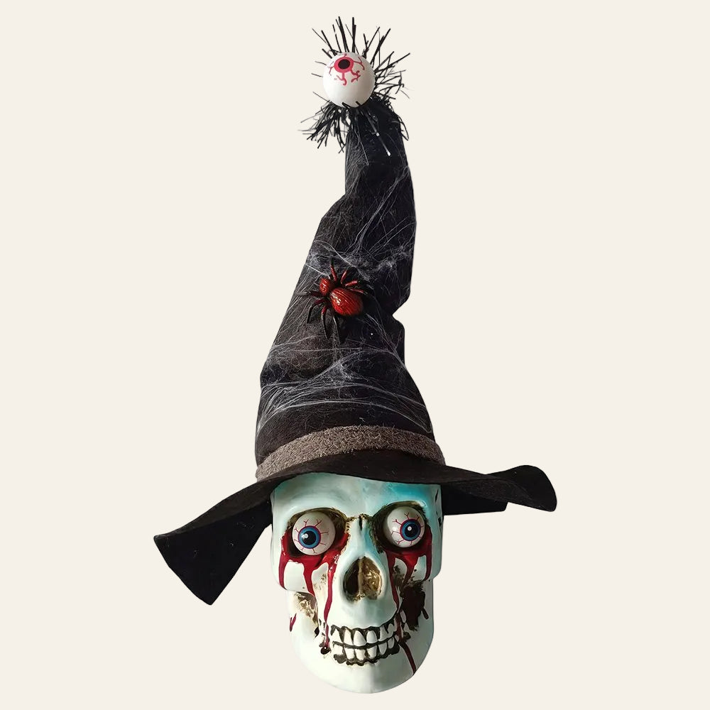 Decoratieve schedel in wit kunststof met een zwarte heksenhoed op met een dikke rode spin en vanboven op de punt van de hoed hangt een oog voor halloween als productiefoto