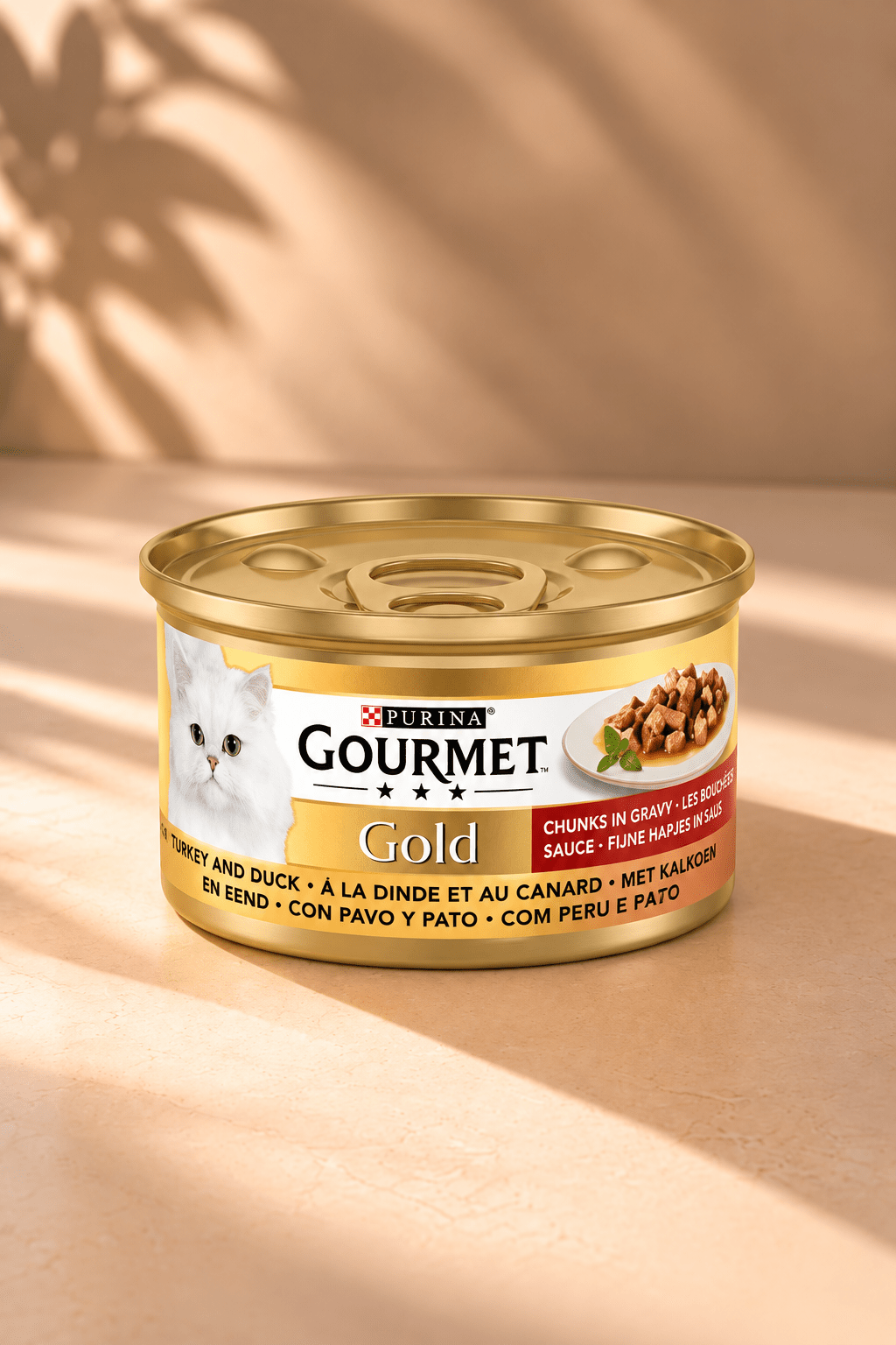Gourmet Gold fijne hapjes met kalkoen en eend in saus, goudkleurig kattenvoerblik van 85 gram op warme beige achtergrond met bladschaduw.